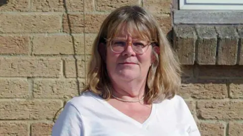 BBC Headshot of Julie, standing outside in front of a pale brick wall. She has straight shoulder-length blonde hair with a fringe and has clear-framed glasses. She is wearing a white v-neck t-shirt and a pearl necklace. 