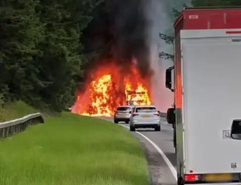 Aaron Wallen A lorry on fire taken from the side of a road. Vehicles are stopped behind the lorry