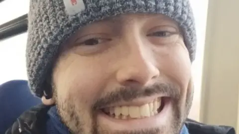 Tyler Foyle A close-up selfie of a man who appears to be sitting on public transport. He is wearing a grey beanie and smiling at the camera.