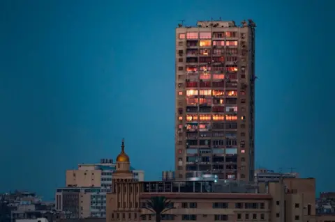 AFP A tall tower block stands next to a domed mosque in a built-up part of the city of Cairo. The sky is a dark blue and the tower's windows are illuminated by the sun in a bright orange. Egypt - Wednesday 20 February 2019