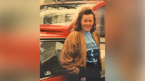 An older picture of Jemma Williamson, a young girl in a brown jacket with a blue T-shirt with the letters SEB and the number '45' visible, has her hands in her pockets and is leaning against a red car which has an open boot