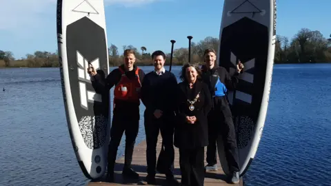 LDRS Three man and a woman stand on a pier with water behind them. Two of the men are wearing life jackets and holding stand-up paddle boards. The woman is wearing a medal.