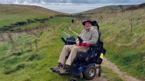 Julie Heath A man in a powered wheelchair parked to one side of a track running down through a field towards the sea. He is wearing a hat and looking towards the camera