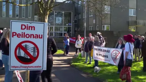 Protestors with placards outside court buildings on grass in sunny weather. The placards show pylons in a traffic sign style symbol with a red line crossed over them and say Dim Peilonau (no pylons)