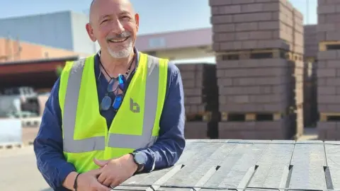 British Recycled Plastic A man in a blue shir tand yellow hi-vis leans on some recycled plasic planks in a builder's yard 