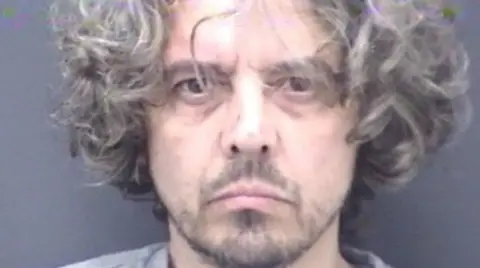 Photograph of Ben Atkins, Dorset Constable, in police custody, with curly gray hair above his ears and unshaven. She is wearing a gray top and standing in front of a dark gray wall.