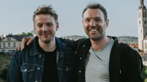 Two men stand, smiling, with a view of a traditional European city spreading out behind them. The man on the right, wearing a black hoodie over a grey t-shirt, drapes his right arm over the shoulder of the other man, who's wearing a blue denim jacket over a black t-shirt.