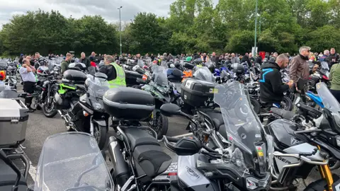 Dozens of motorbikes parked up. In the background are bikers, many dressed in leathers, and other people