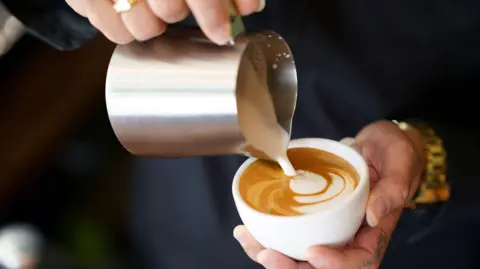 A person is pictured pouring milk into coffee cup