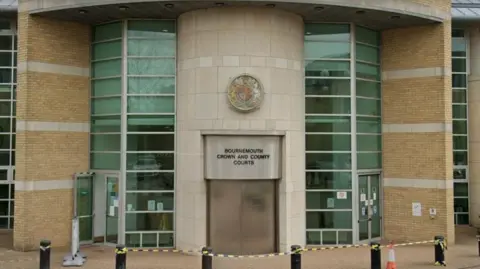 The front of Bournemouth Crown Court. A sign above a large double door at the front of the building reads "Bournemouth Crown and County Courts". Either side of the door are tall strips of windows. In front of the building there is a set of bollards. 