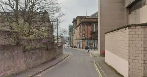 Google A short lane with walls on either side and a red brick building in the background