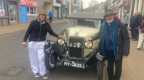 Justin Dealey/BBC Two men standing either side of the bonnet of a vintage car with each having one hand on top of a headlight. The car is parked on the road in a high street. The man on the left is wearing a leather furry hat and the man on the right is wearing a bowler hat and suit with a coat and scarf. There are people on the pavement in the background.