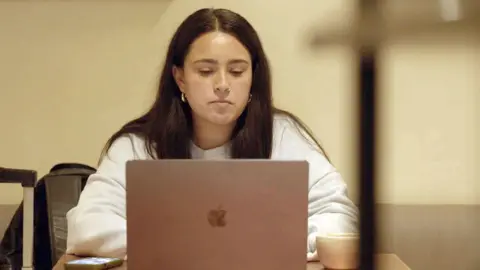 Maya, a woman with long brown hair, a nose ring and earrings, wearing a light-coloured sweatshirt, sits in front of a MacBook in the hospital records office with an iPhone on one side and a cup of coffee on the other.