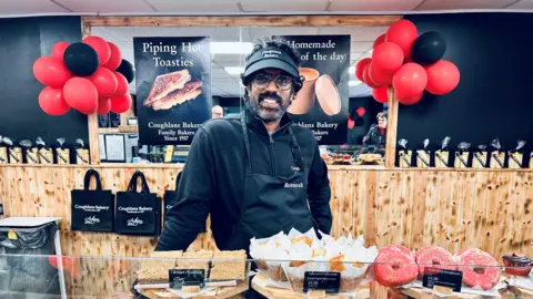 Claudia Sermbezis/ BBC Romesh stood behind a counter of baked goods. He is wearing an apron and black cap and is smiling directly at the camera. There are red and black balloons behind him