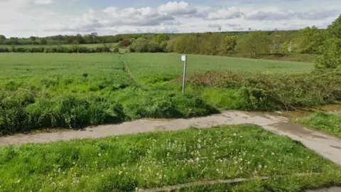 A field covered in grass, with a footpath and the front of the image. The field has a public right of way cutting across it, with trees and hedges surrounding the land. Three sets of floodlights can be seen in the distance. 