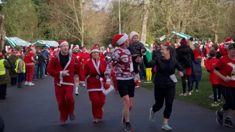 A man and woman running in the Santa Dash with the man carrying their daughter