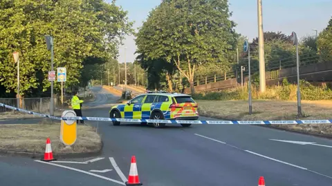 A road has been blocked off with small cones and blue and white police tape, which is stretched across the image. A police car is parked in the middle of the road and an officer is standing next to it.
