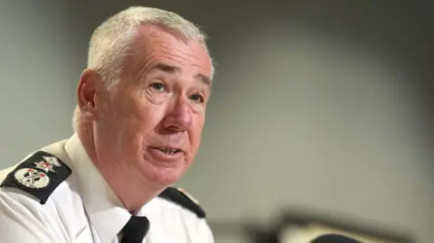 PA Media Jon Boutcher has short white/grey hair and is wearing a white police shirt and black tie