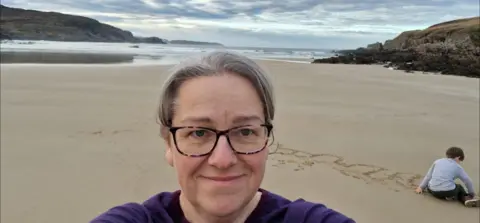 Sally Crowe Sally smiles as she takes a landscape selfie on a stunning beach. Her young son is playing in the sand behind her, writing letters in the sand.