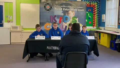 A man in grey clothes sits in a classroom facing a table where a number of schoolchildren, one girl and three boys are sitting. There are several signs on the table with PSNI symbols which say 'Kids Court'.