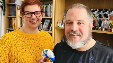 Golden Cat Studios Emily Garside with short red hair and glasses, wearing a mustard yellow knit jumper, stood next to a man with short grey hair and a beard. He is holding a small piece of Doctor Who merchandise and they are stood in front of bookcases full of other fandom merchandise.