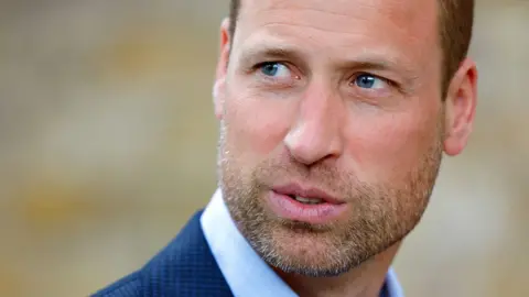 Prince William, head and shoulders photo as he looks away from camera wearing a suit and blue shirt with an out-of-focus beige background, pictured in London in September 2024.