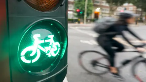 A green traffic light signal illuminates a bicycle on a traffic light. In the blurred backshot is a cyclist on a road.