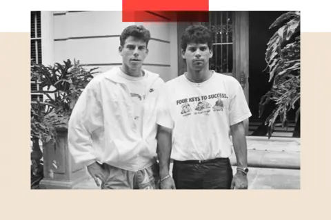 Getty Images The Menendez brothers, Erik on the left and Lyle on the right, pose for a photo. Erik wears a graphic T-shirt partly covered by a light jacket, with one hand in his trouser pocket. Lyle sports a similar T-shirt, black trousers, and a watch on his right wrist. They stand in front of a gate