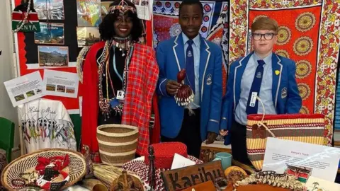 Handout Pupils and a member of staff in a display at the school's multi-cultural day. She is wearing a red scarf and lots of African jewellery and the school pupils are both boys wearing a blue uniform. The boy in the middle is holding a brown shaker. The stall they stand at is full of African accessories and wooden ornaments and jewellery and rugs and clothes and pictures.