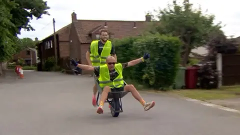 Two men are competing in a wheelbarrow race, both in yellow hi-vis jackets. One man pushes another man who sits in the wheelbarrow with his arms outstretched wide, and wears black sunglasses and gloves. He is bald. The man pushing has brown hair, and his tongue out towards the camera. Behind the men is a residential area, with brown brick houses, green shrubbery, wheelie bins and orange cones surrounding them. The floor beneath them is a light grey concrete.