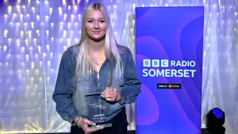 Ruby Andrews with long blonde hair, wearing a blue denim zip-up jacket and holding her award in her hands while smiling at the camera. There is a blue BBC Radio Somerset banner screen to her left. 