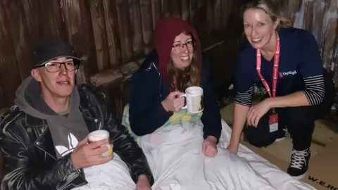 A Threshold member handing out warm drinks and talking to people in Swindon. The threshold worker is wearing a charity top and crouching down, smiling. A woman and a man are under a duvet holding hot drinks and smiling. The woman is wearing a red hoody and glasses and the man is wearing a dark cap and glasses. 
