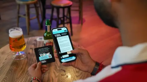 Marston's A man is holding a beer mat in one hand and a mobile phone in the other above a table with alcohol on it. Part of the side of his head, including a beard, is visible.