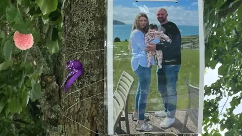 A photograph of mum Gwen, dad Rob and baby Mabli is tied to a tree. The mum and dad are smiling at the camera holding baby Mabli together in their arms, with the sea in the background. 