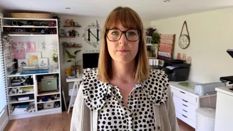 Becki Bowden / BBC Natalie Ramsell-Lee. She has short brown hair, a fringe and wears glasses. She is also wearing a black and white polkadot blouse. She is sat her desk, with one of her designs on the screen, and is smiling at the camera.