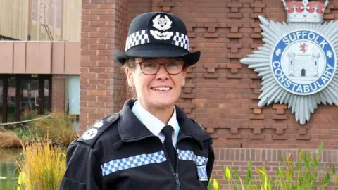 Suffolk Constabulary Rachel Kearton standing outside Suffolk Constabulary's HQ. Rachel is wearing a navy police hat with blue and white trim, a navy police zip-up jacket over a white shirt and glasses. She is smiling at the camera.