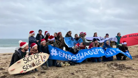 A group of people are holding banners and signs on a beach, with the sea behind them. One sign - on a surfboard - reads sick of sewage.