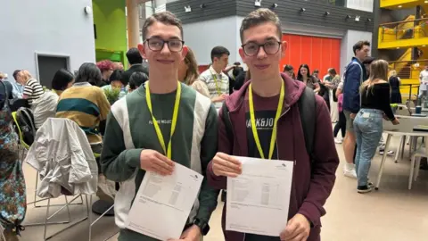 BBC Identical twins, both with glasses, one wearing a green jumper and one in a red jacket. Both are holding up their A-level results. 