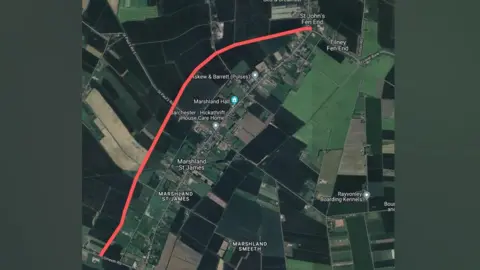 Google An aerial satellite style map showing the footpath highlighted in red, stretching from the top of the map at St John's Fen End down to Smeeth Road. The fields are dark and light green.