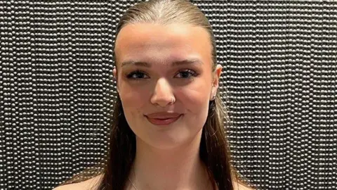 A close-up shot of Ava who has her long brown hair tied up in a ponytail and is wearing a nose ring and silver necklace