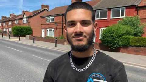 Young man with a beard and dark hair, eyes and skin, wearing a black t-shirt and silver chain, standing on a street in Maltby, Rotherham with a pavement and red brick houses and a hedge behind.