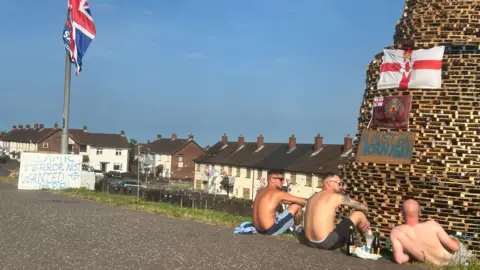 Three topless men sit in front of an unlit bonfire, with bottles of wine beside them. A Union Jack hangs limp from a post near them. 