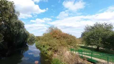 Google A river reflecting clouds is visible to the left, flanked by trees and hedges. A path edged with green railings is on the right with a tree in the background.