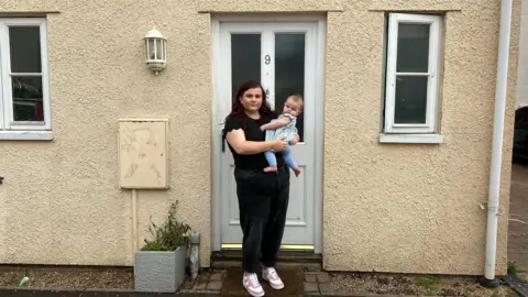 a woman dressed in black holding her baby on her hip standing outside the white front door of a terraced cream-painted house