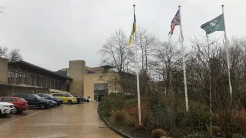 Three flags, including the Union Jack and the logo of Surrey County Council, fly outside the council's headquarters at Woodhatch Place in Reigate.