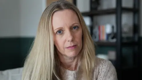 John Fairhall/BBC A blonde woman in her forties looking at the camera with a neutral expression. She is wearing a knitted jumper. There is a black coloured cabinet behind her.