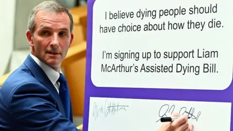 Getty Images A man with dark greying hair writes his signature on a board pledging his support for assisted dying. He is wearing a blue suit, white shirt and blue tie. His head is turned to the side as he writes on the board with his right hand.