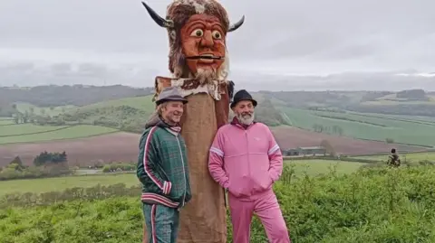 Vic Irvine Chesney Hawkes and Vic Irvine stand either side of the Dorset Ooser. They are surrounded by countryside and up on a hill. Chesney stands to the left, wearing a green and red tartan print tracksuit, smiling as he looks at a camera to the right of the frame, and has a brown hooded puffer jacket on under his tracksuit with a grey trilby style hat. Vic Irvine stands to the right, he has a grey beard and wears a black trilby hat and bright pink tracksuit set. In the middle of the pair is the Dorset Ooser, a wooden head from 19th century folk culture. The head has a humanoid face with horns, a beard, and a hinged jaw which allows the mouth to open and close. 