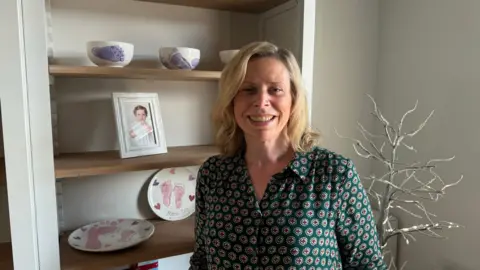 BBC Mother Nikki smiling at the camera in front of a picture of her daughter when she was a toddler. There are plates on the shelf behind Nikki with prints of her daughter's feet.