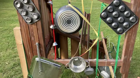 Guy Campbell/BBC An array of kitchen utensils and frying pans are hanging from a wooden frame in playground where children can create sounds and rhythms 